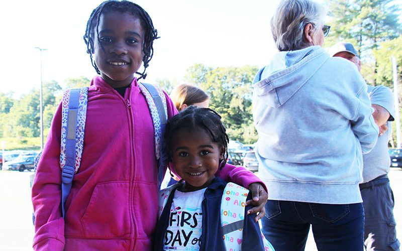 Two elementary students stand together, with arms around each other. They are smiling. The smaller child is wearing a shirt that says First Day of Pre-K