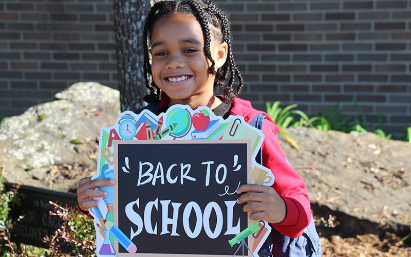 A girl smiles and holds up a blackboard sign that says Back to School.