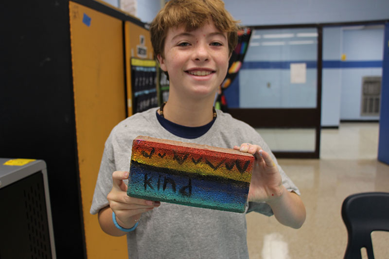 A sixth-grade kid smiles and holds up a brick they painted in rainbow colors.