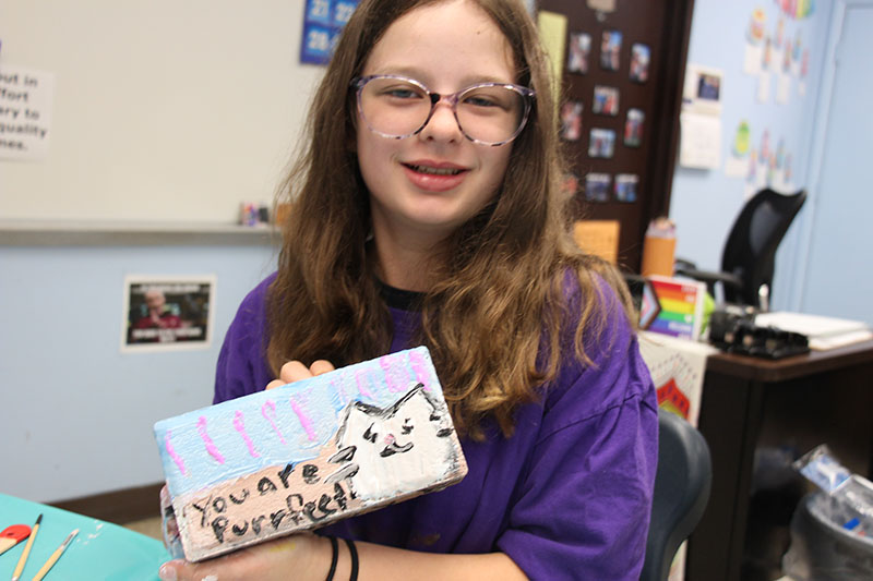 A girl withlong brown hair wearing glasses and a purple shirt, holds up a brick she painted that has a picture of a white cat and the words you are purrfect.