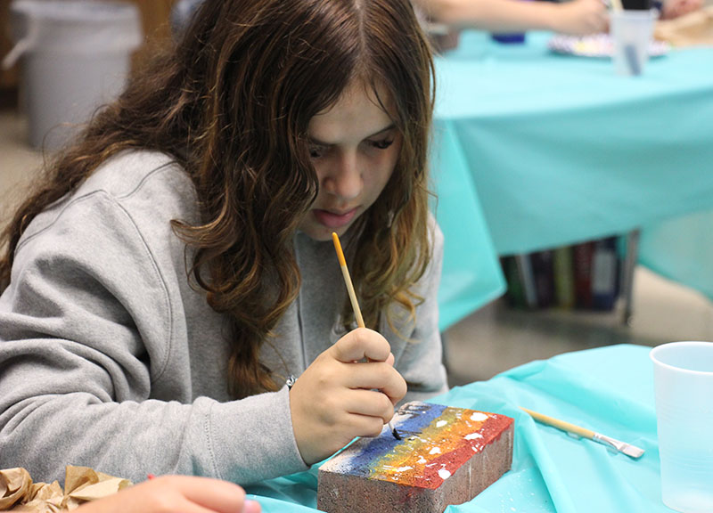 A sixth-grade girl with long brown hair paints a brick.