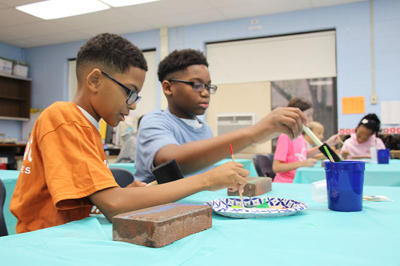 Two sixth-grade boys sit at a table painting bricks.