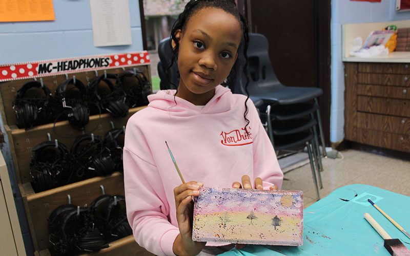 A sixth-grade girl in a pink sweatshirt smiles as she holds up a brick she painted for the kindness garden.