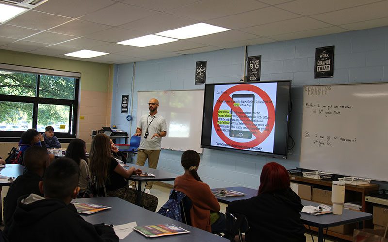 A man stands in front of a class talking to middle school students. On a display is a cell phone with a large red line through it.