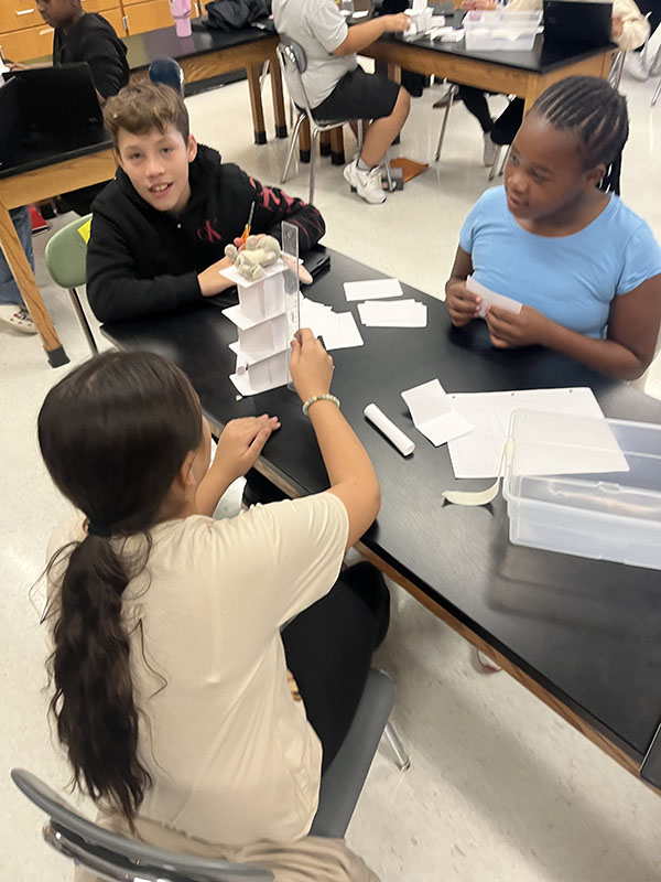 Three sixth grade students work at a table building an index card tower.