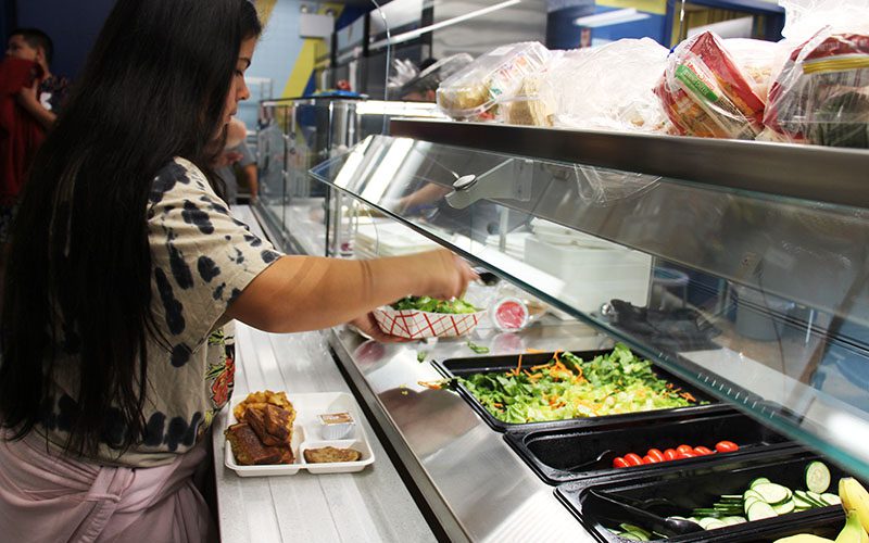 A girl with long black hair stands on a lunch line and putt salad into a dish.