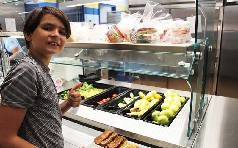 A middle school boy gives a thumbs up holding a tray of french toast sticks. a salad bar is in the background.