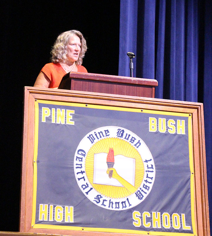 A woman with shoulder length blonde hair stands at a podium and speaks. In front is a large framed banner that says Pine Bush High School.