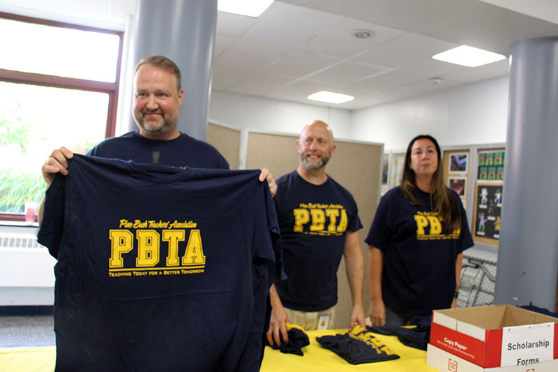 A man holds up a navy blue tshirt that says PBTA. Two others look on smiling.