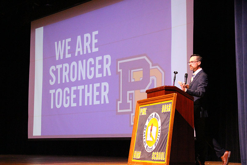 A man stands at a podium speaking. Behind him is a large screen that says We are stronger together.