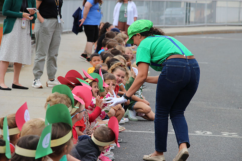 A woman in a green hat and shirt dressed as the cartoon character Luigi, goes along high fiving students who are sitting on a curb.