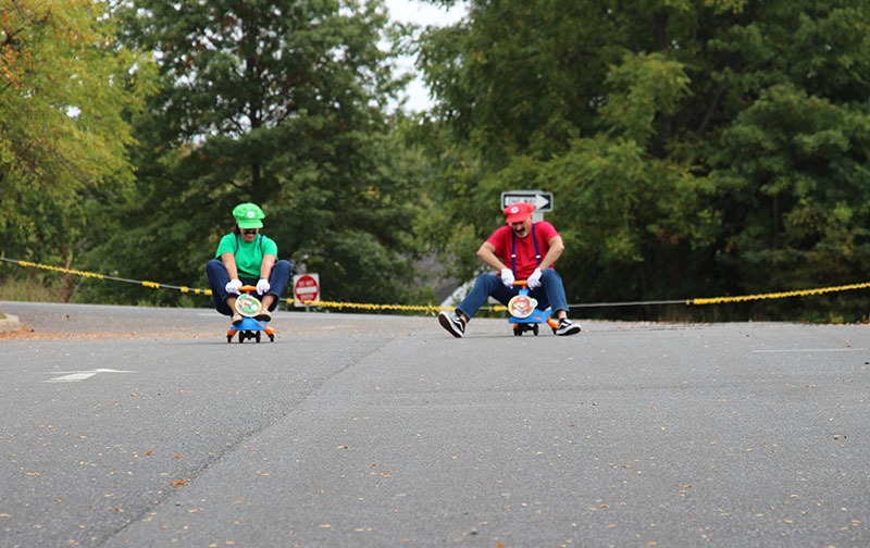 Two adults dressed as Luigi and Mario sit on little scooters and race down the street.