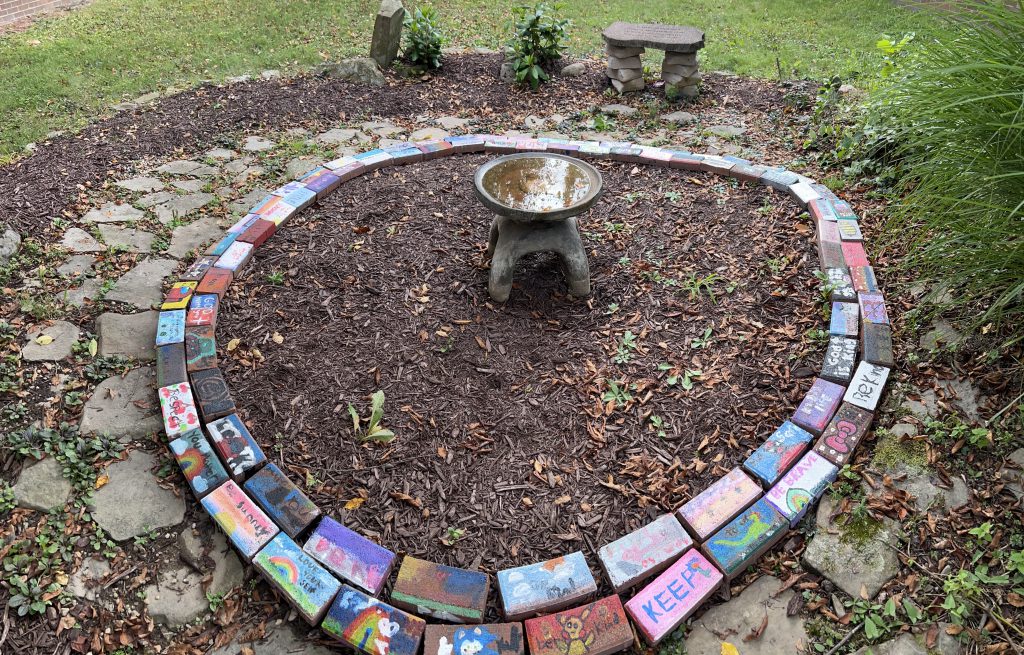 An area under a tree filled with mulch and two large rings of painted bricks. In the center is a metal stand.