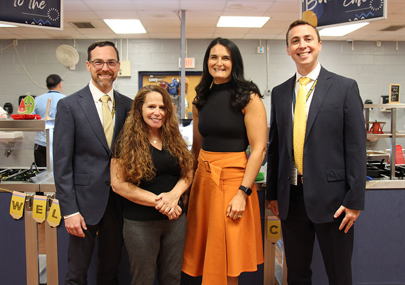 Four adults stand together, men on either end and two women in the center. They are all smiling.