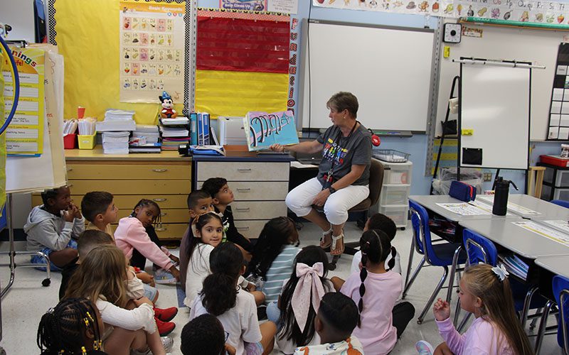 A woman sits on a chair and reads from a book to a group of second grade students who are sitting on the floor.