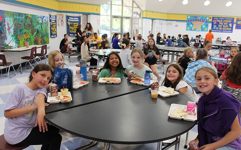 Six elementary girls smile as they sit around a lunch table. Each has a tray of food in front of them.