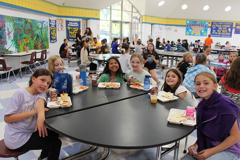 Six elementary girls smile as they sit around a lunch table. Each has a tray of food in front of them.