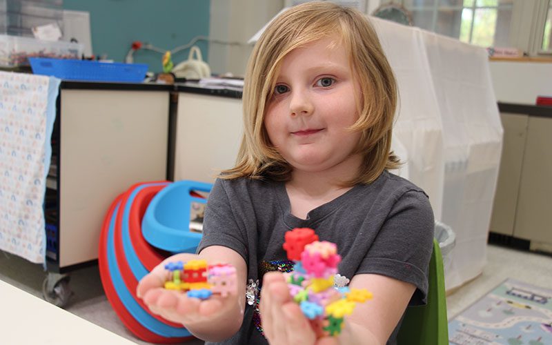 A kindergarten student with blonde hair holds out something she made with little blocks