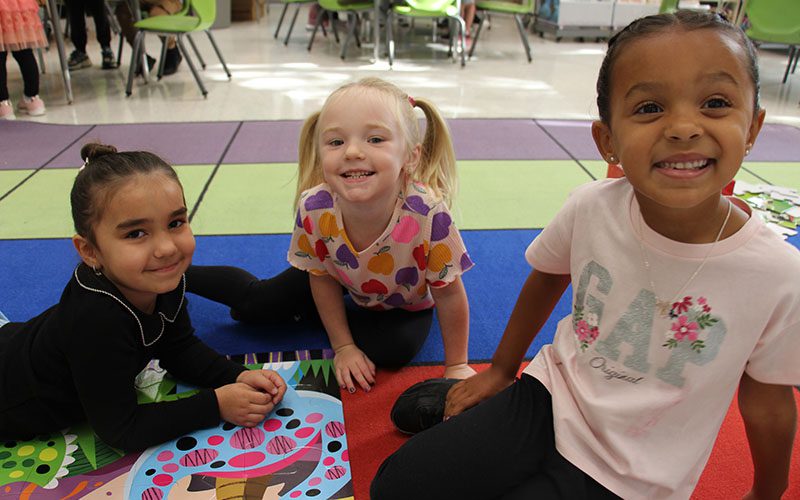 Three little girls sit on a colorful rub and smile.