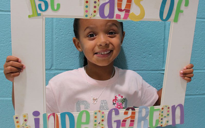 A little girl smiles as she holds a photo frame that says 1st Days of Kindergarten