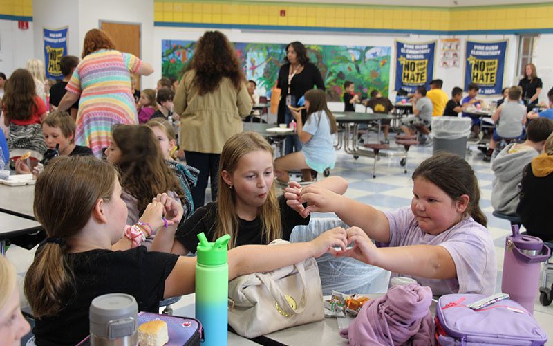 Three girls sit at a lunch table and join hands to form hearts.