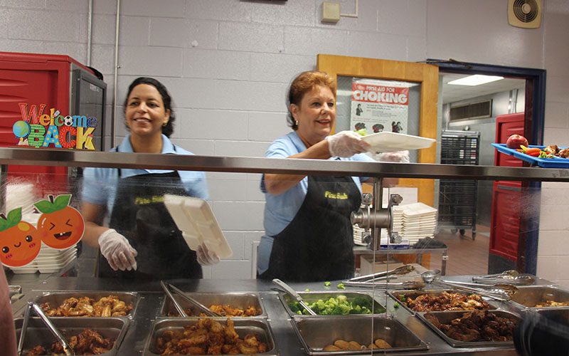 PBHS Food services handing tray Two women wearing blue shirts and black aprons hand food trays to students.
