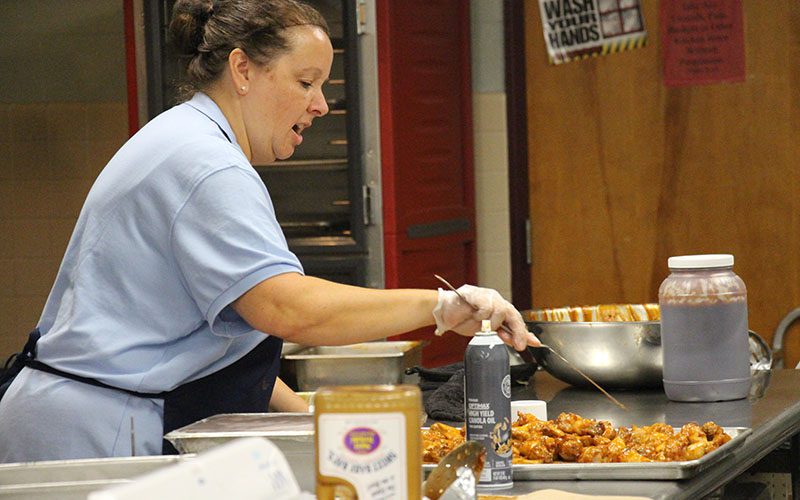 PBHS Food services making wings A woman wearing a blue shirt and apron stirs wings on a large baking sheet.