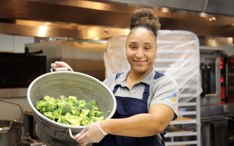 PBHS Food services worker with broccoli A woman smiles as she shows a very large colander filled with broccoli. She is wearing a gray shirt and blue apron.