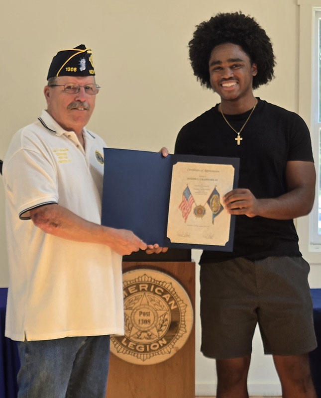 A high school young man smiles as he accepts a certificate from a man on the left, who is wearing a white shirt and blue American Legion cap.