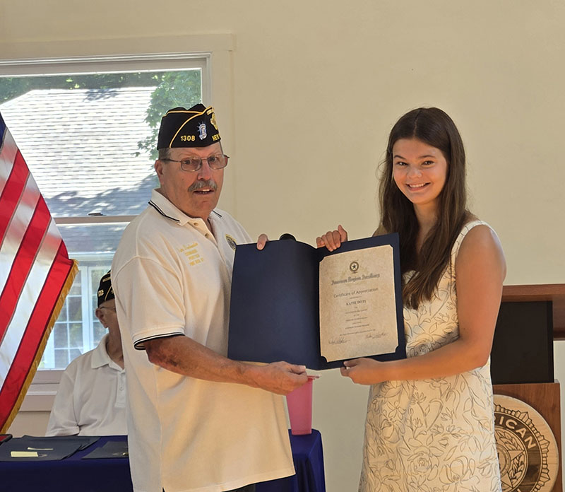 A high school young woman smiles as she accepts a certificate from a man on the left, who is wearing a white shirt and blue American Legion cap.