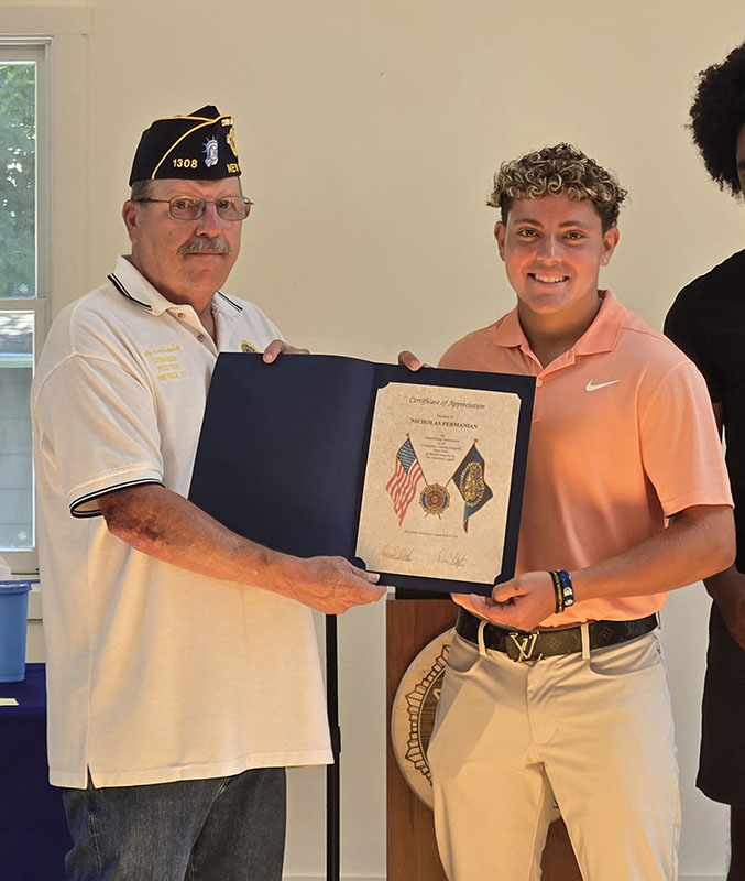 A high school young man smiles as he accepts a certificate from a man on the left, who is wearing a white shirt and blue American Legion cap.