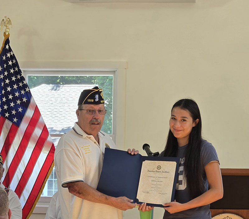 A high school young woman smiles as she accepts a certificate from a man on the left, who is wearing a white shirt and blue American Legion cap.