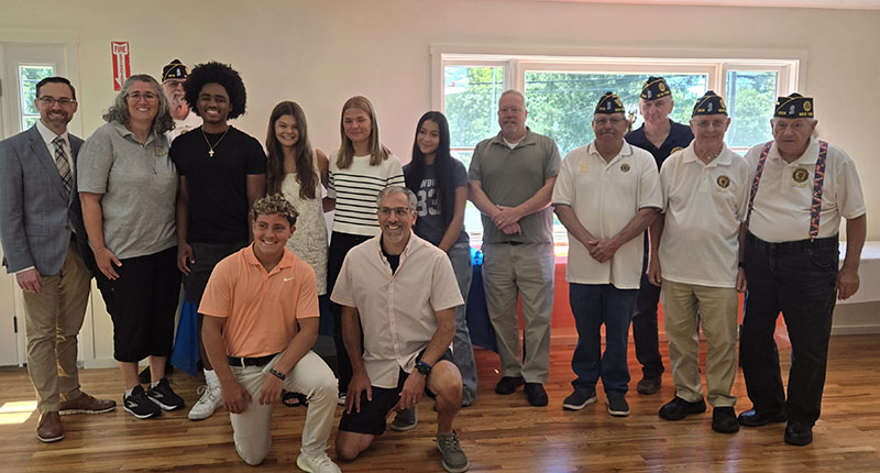 A group of five high school students and eight adults stand together in two rows. All are smiling and some of the adults have American Legion caps on.