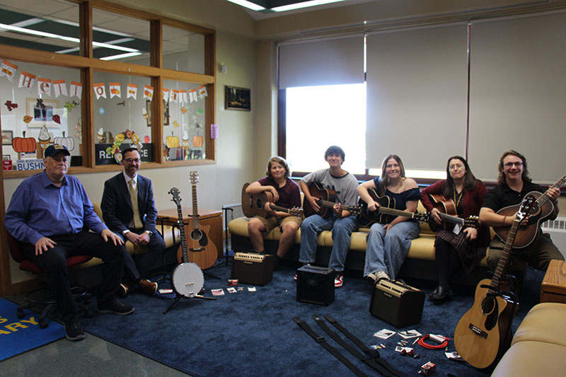 A group of four high school kids and an adult woman sit on a couch holding guitars. Next to them two men sit and smile. There is other guitar equipment on the floor.