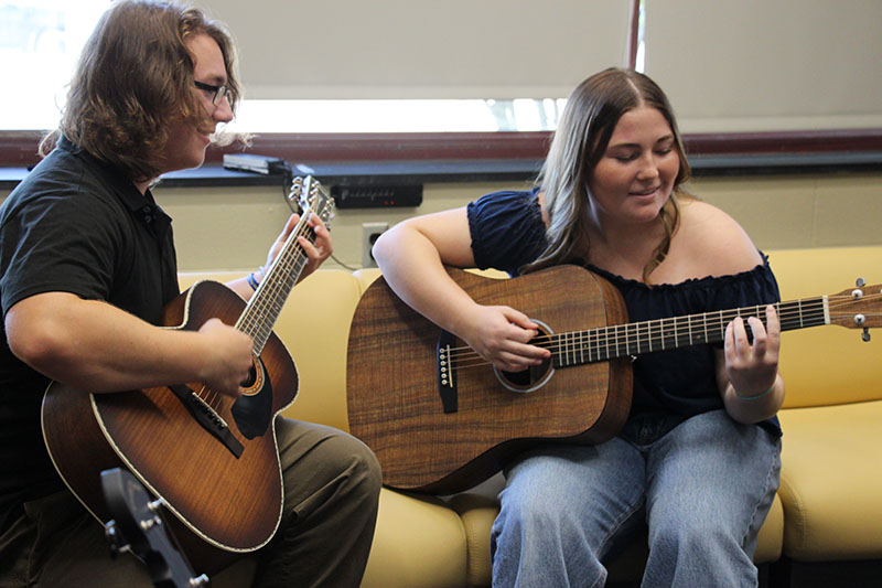 Two high school kids sit on a couch playing acoustic guitars.