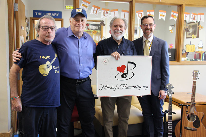 Four men stand and smile. One holds a white sign that says Music for Humanity.
