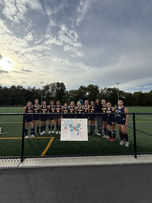 A group of girls wearing navy blue field hockey uniforms stands around a poster that says Morgan's Message.