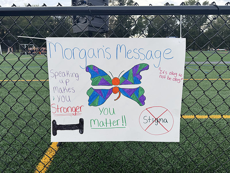 A poster hanging on a chain-link fence. It says Morgan's message with a butterfly in the center. It also says Speaking up makes you stronger. You Matter! It's okay to not be okay. and a red x over the word stigma.
