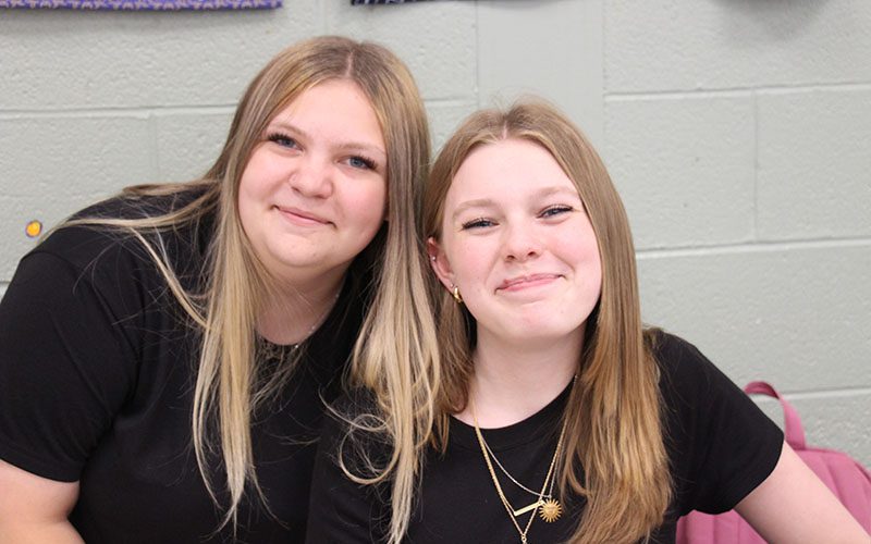 Two high school girls, both with long blonde hair wearing black shirts, smile.