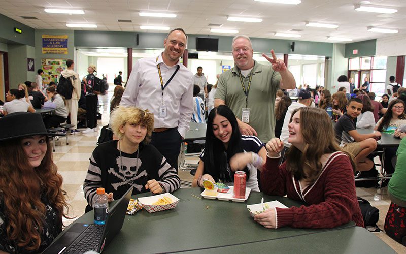 Four high school students sit at a table with their lunch in front of them. They are laughing. Two men stand behind them smiling. The man on the right is giving the peace sign.