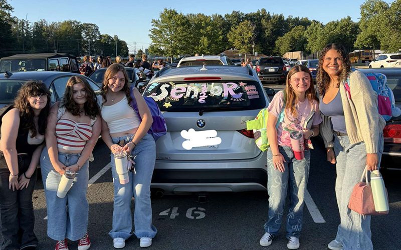 A group of of five girls smile. They are standing around a car that says Seniors 2026 on the back window.