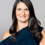 A woman with long dark hair, wearing a dark blue dress, smiles.