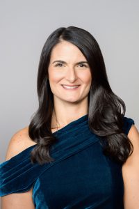 A woman with long dark hair, wearing a dark blue dress, smiles.