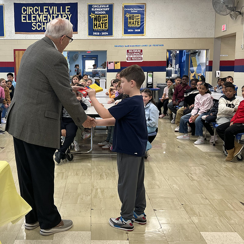 A man on the left shakes hands with a third-grade boy who takes a dictionary in his other hand. A large group of kids sit at tables watching.