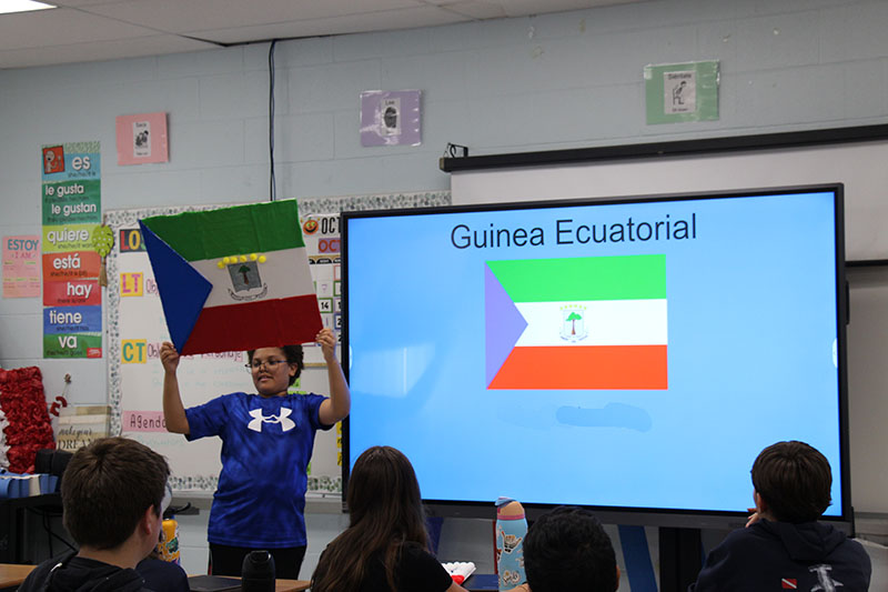 A seventh-grade boy holds up the flag of Guinea Ecuatorial. It is also displayed on a screen next to him.