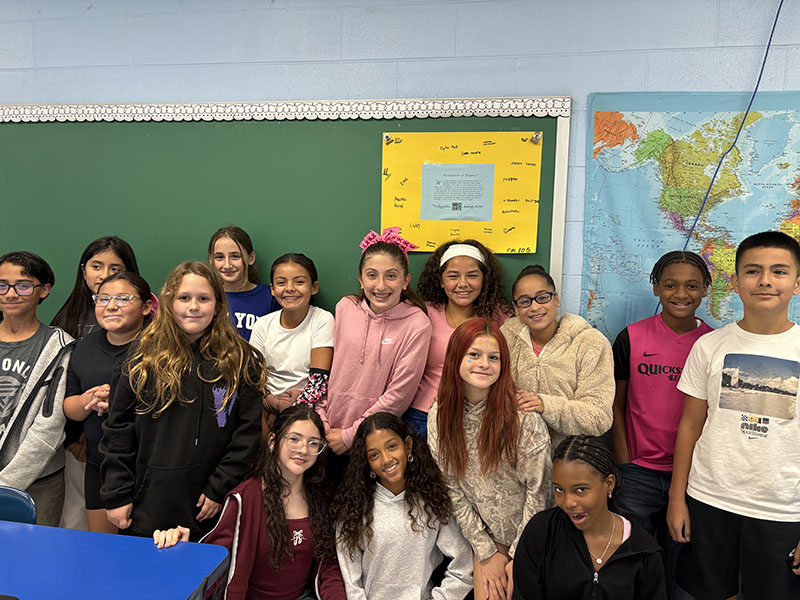 A group of middle school students stand together smiling. Behind them is a poster with their signatures on it.