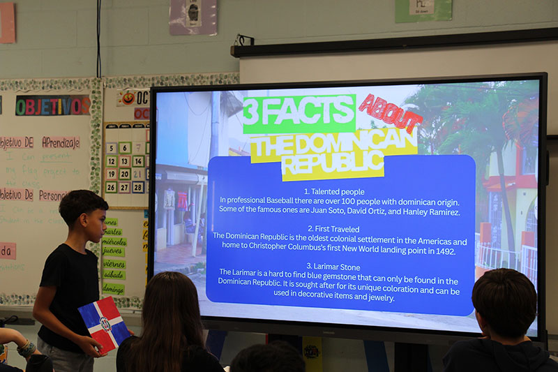 A seventh-grade students stands next to a board that has a colorful presentation on it called 3 Facts about the Dominical Republic.