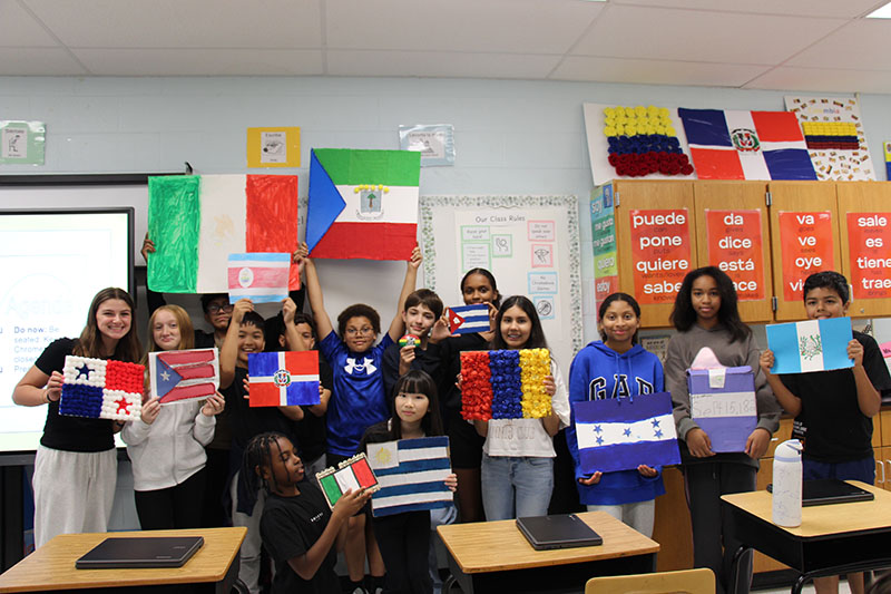 A group of a dozen seventh-graders stand together holding the flags they made of Spanish-speaking countries from around the world.