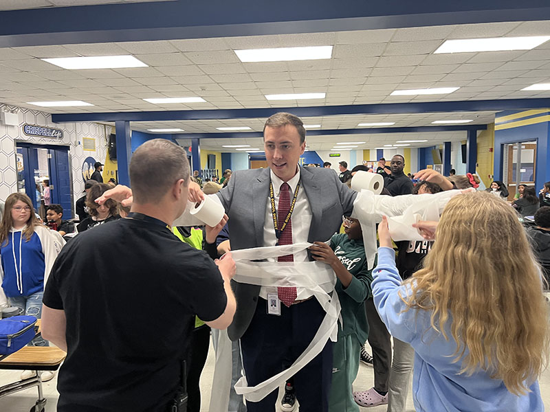 A man in a suit and tie smiles as kids around him start wrapping him in white toilet paper.