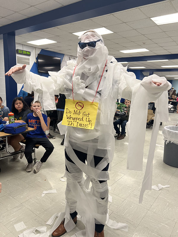 A man is fully wrapped in white toilet paper and is wearing black sunglasses. He has a yellow sign around his neck that says Do no get wrapped up in drugs.